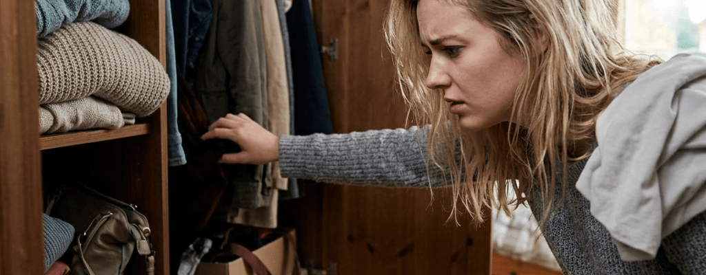 Woman with blonde hair looking frustrated while searching through disorganized clothes in a wooden wardrobe.